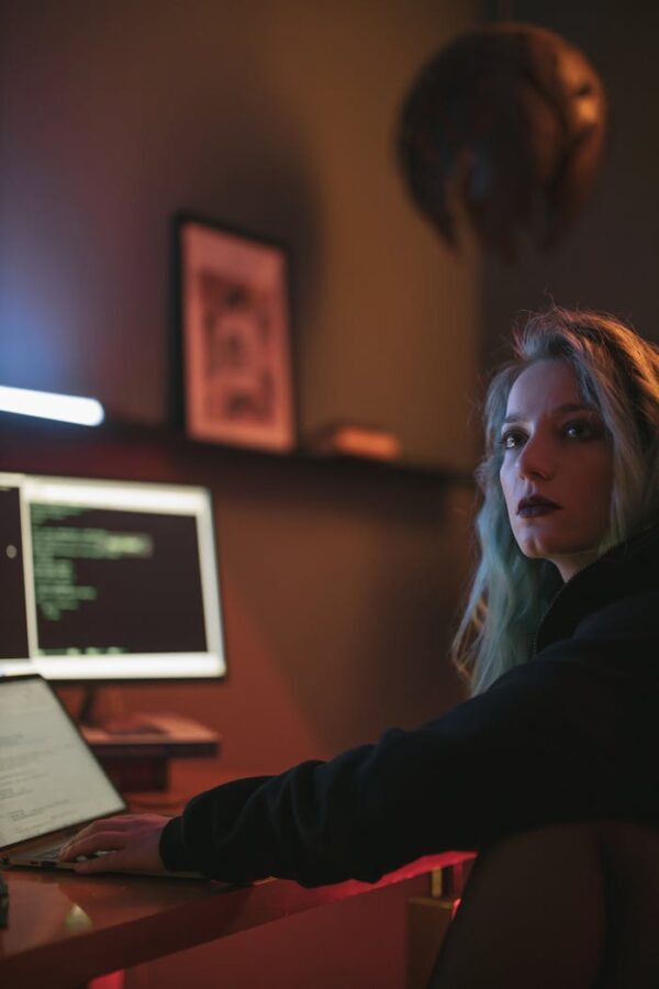 A woman with colorful hair works intently at her computer in a dimly lit room, coding on a monitor.