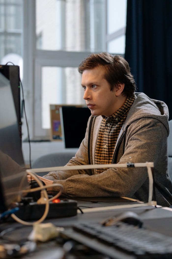 Young programmer in hoodie focused on coding at a desk in a modern office setting.